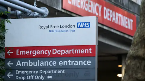 Getty Images A white sign outside the hospital reads "Royal Free London", with a red strip underneath saying "Emergency Department" in white lettering, and grey strips below saying "Ambulance entrance" and "Emergency Department Drop Off Only" in white lettering, with a white symbol of a car. In the background a red sign with white lettering marks the Emergency Department entrance