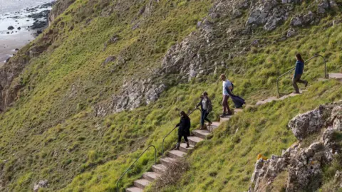 National Trust /James Dobson Four people walking down the steps at Brean Down, which are carved into the sloping hillside with a green metal handrail. At the bottom of the grassy hill is the beach full of craggy rocks at low tide.