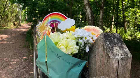 Jessica Lane/BBC A bouquet of colourful flowers tied to a gate. There is a dusty path and woodland behind it.
