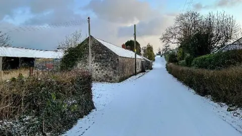 Weather watcher Ian A lane is covered in white snow and runs from top to bottom of the frame. There is a dark bricked cottage to the left of the lane and a green and brown hedge to the right, which runs parallel to the road. There is a grey barn behind the cottage The sky is blue and partially cloudy.