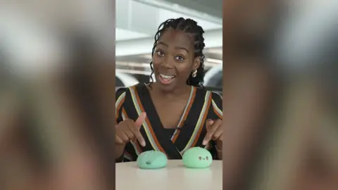 A woman with dark hair pulled back from her face points at two similar squishy toys on the desk in front of her