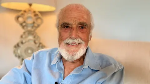 Alex Dunlop/BBC Chris Crowther looks directly at the camera and smiles. He has white hair and a beard and is wearing a light blue open-necked shirt. Behind him is an ornate lamp.