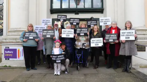 A group of people standing in front of Sudbury Library, holding signs saying "Don't wreck our libraries".