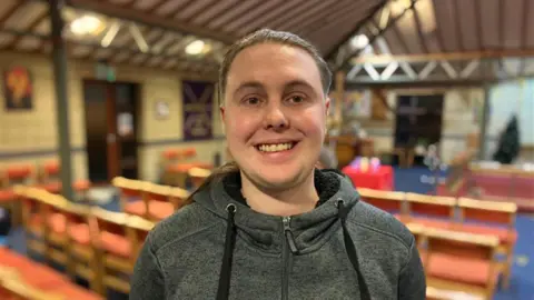 James Tomkinson is wearing a grey hoodie and is smiling at the camera, with the rehearsal space in the background. There are rows of seats in what looks like a church hall