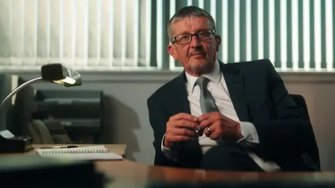 Sean De Francesco/Firecrest Films Alan Smith sitting behind a desk, with a lamp to his left, in front of a window with vertical blinds. He is wearing glasses and is dressed in a black suit with a white shirt and a green tie.