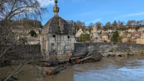 Bradford-on-Avon Town Bridge in Bradford-on-Avon with the river level right up to the parapet and a large tree trunk which has been swept down the river and is jammed against the bridge