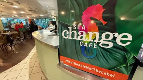 A Sea Change Cafe sign displayed at the counter. It reads: #SeeTheAbleNotTheLabel. In the background are tables and chairs, as well as some people.