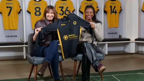 Wolves Foundation Two women are sat on chairs in a football changing room surrounded by orange football shirts. Between them they are holding a long sleeve black football shirt 