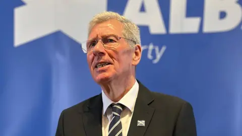 BBC Kenny Macaskill wears a dark suit and striped tie, glasses and has short white hair