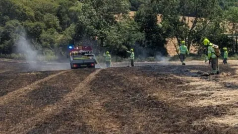 ECFRS Five firefighters dampen down flames in a large field. They are wearing yellow high-vis uniform. The grass is charred and there are green trees in the background.