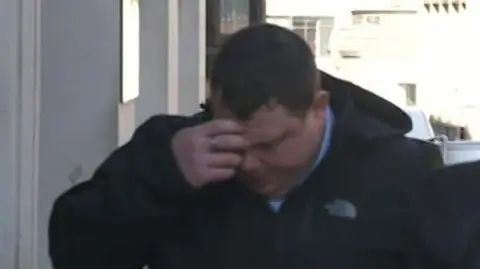 Still taken from video of a man walking into court. He is wearing a black North Face jacket and is holding his hand to his head.