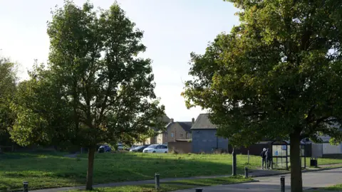 A tree-lined path in Cambourne, in the distance are houses and there are two people waiting beside a bus stop.