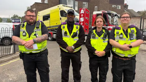 Amy Holmes/BBC Four high-visibility uniformed council personnel pose in front of recovery vehicles and a former emergency response vehicle that is about to be towed away from an urban roadside setting.
