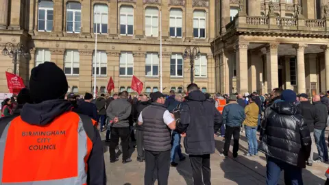 People outside Birmingham council house, pictured from behind. The person nearest the camera has a high-vis orange jacket which says 'Birmingham City Council' on the back. Several red 'Unite' union flags can be seen and in the background is the council's cream building which has an entrance of pillars and and a balcony. 