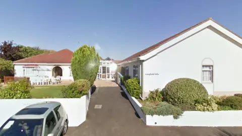 Longfield Villa is printed on the wall of a single storey building set on a  drive which leads to two other single storey buildings, one to the left and one behind. In the garden there are many plastic chairs set around garden tables and sun shades. There is a car parked off the road.