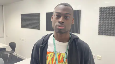 Tobi has very short black hair and black facial stubble. He is wearing a white t-shirt with a graphic comic book type design on it. He has a light blue lanyard around his neck and is wearing a dark navy zip hoodie
