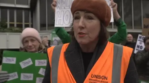 LDRS/Oliver Leader de Saxe A woman wearing a brown beret, an orange hi-vis jacket and a black jumper. She is standing next to a group of protesters outside a local council building.