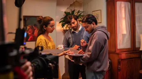 Pinder Productions, Geppetto Pictures, Kai Campbell The producer, director and crew member stood over a script making changes, behind the scenes and in front of a lens of a big black camera. They are stood in a house which has a painting on the wall and a plant behind them.