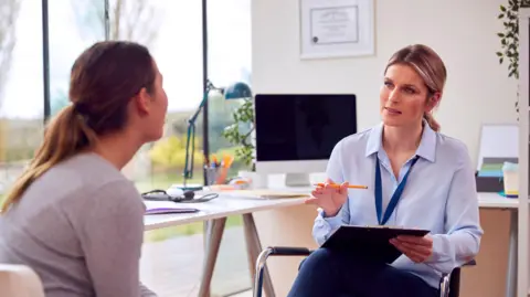 A female doctor in an office meeting with a female patient. They are sat opposite each other. The doctor has a clipboard in her lap and is holding a pen. Behind the pair are large windows that look out onto a garden.