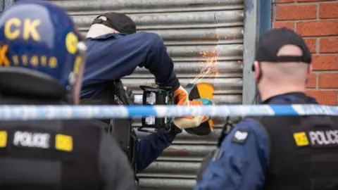 GMP Police are carrying out a raid on the property on Lockett Street. Three police officers are facing towards a metal roller shutter - one is using angle grinder with sparks flying into the air.