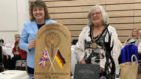 A woman in a baby blue blazer holds a wooden plaque with the United Kingdom and German flags on it with a flame inscribed on it. A woman in a white and black dress with a mayoral chain stands next to her.