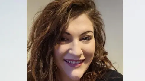 Portrait image of Charlotte Smith looking towards the camera smiling. She has dark brown hair in a side parting, and is wearing purple lipstick and a black T-shirt.