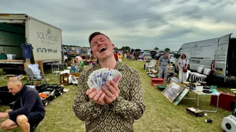 Sonny Green A man standing in the middle of a car boot sale with a wad of cash notes in his hand, his head is tipped back with a look of bliss on his face.