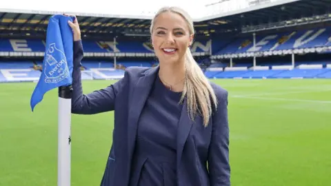 Everton Elle Barnes-Reen is standing pitch side at Goodison Park with her right arm holding the corner flag. She is smiling directly at the camera