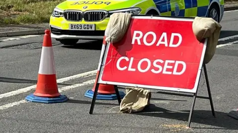 A road closure sign in front of a police car. There are three orange cones around the red sign. 