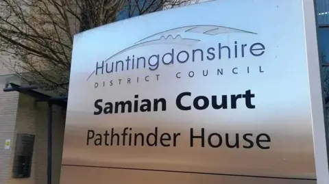 Local Democracy Reporting Service A silver sign is off centre to the right. It has the Huntingdonshire District Council sign at the top and says Samian Court, Pathfinder House below.