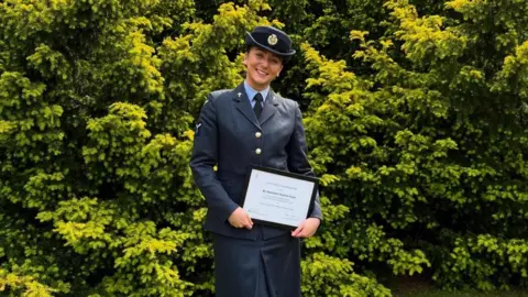 Jess Corps A woman in RAF uniform standing outside smiling and holding a certificate. There is greenery in the background and it is a sunny day.