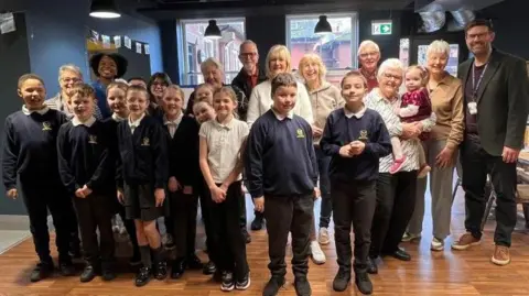 A group of school children, who are wearing their school children, are standing alongside a number of adults who are taking part in the pen pal scheme. They are all smiling at the camera. 