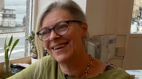A headshot of Phoebe looking to the right of camera and smiling. There is a window to her right and she is wearing a green jumper and a chunky gold necklace and glasses.