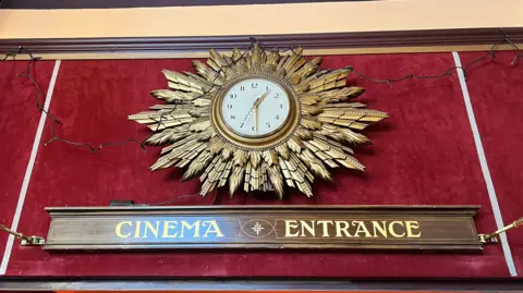 Above a wooden sign with gold lettering saying cinema entrance is a circular clock with gold coloured hands on a white background, with an elaborate golden surround looking like lots of feathers.