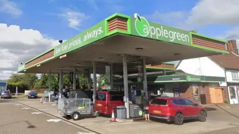 Google Street View image of a petrol station forecourt. There are red cars next to the pumps. Neighbouring houses can be seen. The petrol station is covered by a canopy bearing the words "applegreen" and "low fuel prices, always". 