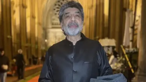 BBC Arif Khan wearing a black silk shirt standing in a cathedral 