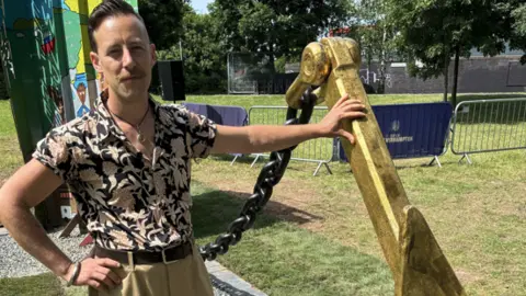 Perry stands by the side of the anchor, resting his hand on it. He has a patterned shirt on and brown trousers.