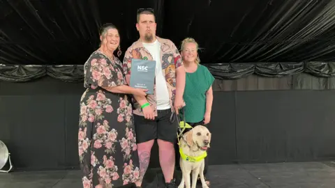 Three people stand on a stage with a guide dog wearing a high-visibility harness. One person holds an National Star College Record of Achievement certificate while posing for a photograph.