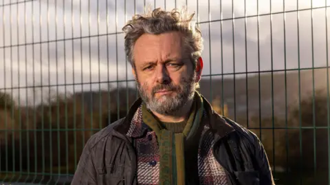BBC/Wall to Wall Media/Stephen Perry. La imagen es un retrato al aire libre, tomado cerca de una valla de seguridad de malla metálica. Michael Sheen posa frente a la cámara, vestido para el clima frío con varias capas de ropa, incluyendo una chaqueta oscura de estilo militar, un abrigo de lana a cuadros y una bufanda verde estampada. La valla ocupa gran parte del fondo, mientras que las colinas y los árboles distantes se ven suavizados por una profundidad de campo reducida. La luz dorada del sol, desde un ángulo bajo, aporta calidez a la escena y resalta las texturas de la bufanda, el abrigo y la valla.