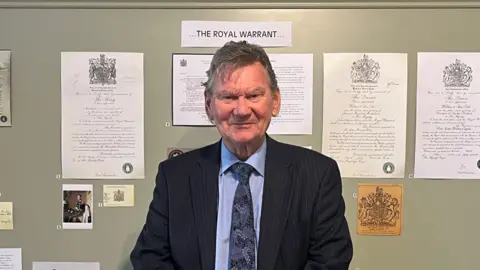 Wilkin & Sons Ltd Man smiling at camera in front of a board titled 'The Royal Warrant'.