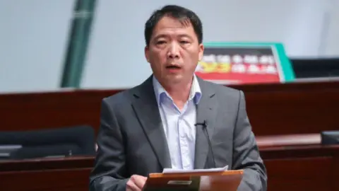 Wu Chi-wai speaking at the legislative council chamber. He's wearing a grey suit and holding sheets of paper.