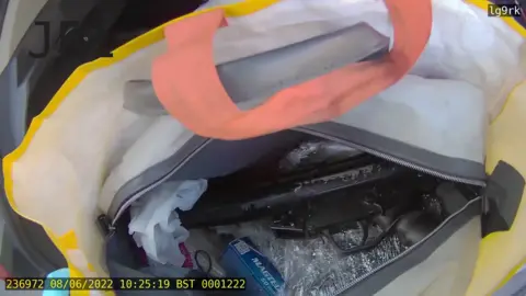 PSNI On overhead view inside a yellow shopping bag with an orange handle.
Inside that bag is another white bag which is unzipped.
Inside it you can see a black gun lying on it's side.
On the bottom left hand side of the picture there is yellow text which gives the date the footage was taken - the 8th of June 2022 - and the time - 10:25 am