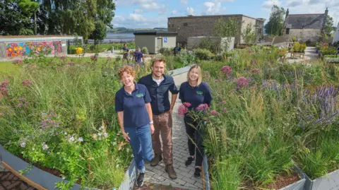Gordon Terris Three people - two women flanking one man - standing in the middle of a Saltire shaped garden. They are all laughing and smiling while surrounded by greenery.