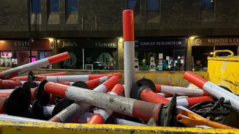 A pile of orange and white bollards in a yellow container in front of a line of shops.