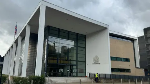 The front of Ipswich Crown Court. A large glass-panelled building with a white white pillars running down one side. It has a large silver sign that reads "Ipswish Crown Court" to its right.  