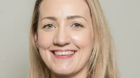 AILG A head shot of Elaine Lynch with long blonde hair. She is smiling and standing in front of a grey background