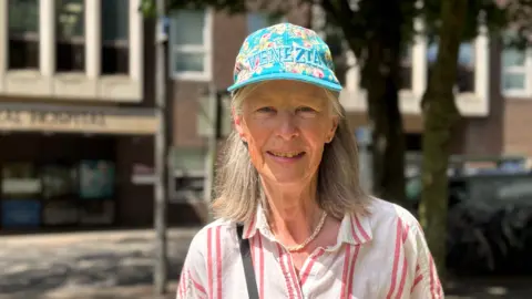 Mary Venturini is standing outside on a sunny day. She is an older woman with light-coloured shoulder-length hair, and is wearing a blue floral and a white and red-striped blouse.
