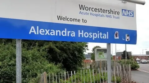 A blue and white hospital sign on a roadside, which reads Worcestershire Acute Hospital NHS Trust Welcome to Alexandra Hospital