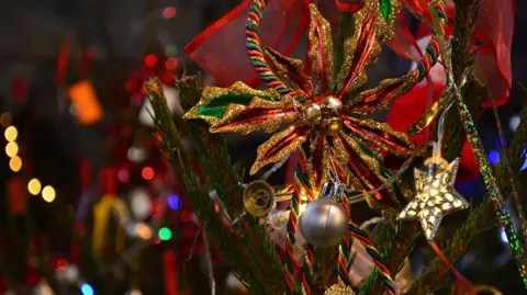 A close-up of a lit-up Christmas tree with baubles, bell and star decorations in front of an out-of-focus background of soft fairy lights