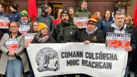 The picture shows 18 people, men and women, standing outside a building holding placards and a banner with a message in Irish on it. Several of the people are wearing woollen hats and all are wearing coats